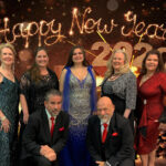 publicity photo for 2019 New years eve with five women in formal dresses standing behind two men wearing black suits with red ties kneeling-