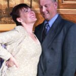 man wearing a gray suit and blue tie smiling and smiling brown haired woman wear a gold sparkling outfit don't dress for dinner