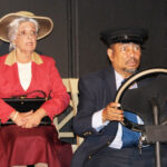 woman sitting on a chair wearing a beige hat, glasses, red jacket, and burgundy skirt holding a black purse with an African American man looking surprised sitting in front of her as if driving a car – driving miss daisy