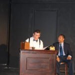 caucasian man wearing a white shirt and green bow tie and glasses sitting behind a wood desk reading a document to an African American man wearing a suite and tie sitting next to the desk