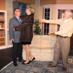 man wearing a suit and tie looking shocked as a tall blond smiling woman wearing a fur coat and black heels embracing him while an older man wearing a yellow shirt and tan slacks looks on smiling-don’t dress for dinner