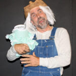 Man with a gray beard and mustache with a head covering with fake white hair, wearing overalls with a white shirt and holding a jar with a green and white checkered dress-mama don’t fly