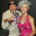 Man in a tan suit holding a champagne glass with is armed crossed with a blond woman wearing a pink dress holding a champagne glass and a twenty dollar bill tucked in her dress-mama don’t fly