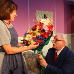 elderly man wearing a navy suit and red tie kneeling and present a large colorful bouquet of flowers to a smiling woman in a checkered dress wearing an apron - sex please, we're sixty