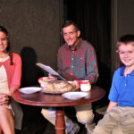 young girl with braids wearing a white dress and pick sweater, man wearing a red and green checked shirt, and young boy wearing a blue shirt and tan slacks sitting around a table set for dinner- christmas pageant