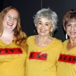 Woman with long red hair and glasses, woman with short gray hair wearing large gold hoop earrings, woman with short brown hair with bright blue eye shadow – all wearing yelling t-shirts spelling out viva las vegas-mama won’t fly