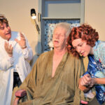 elderly man with eyes closed in a brown robe - red hair woman in a blue floral robe checking his pulse and an upset doctor watching - you should be so lucky