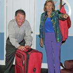 woman wearing blue blouse, purple pants, and plaid jacket pointing her finger while looking at a man who is pushing a red suitcase